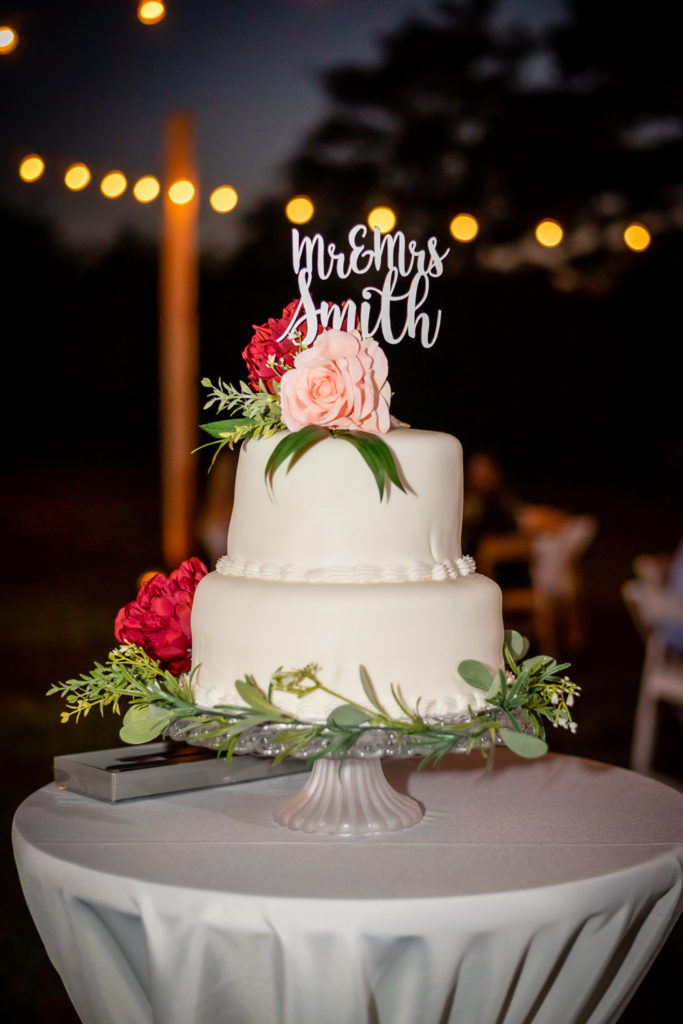 Wedding cake with wooden name topper and pink and red flowers plus greenery at wedding at The Gavi Estate and Barn in Forsyth GA